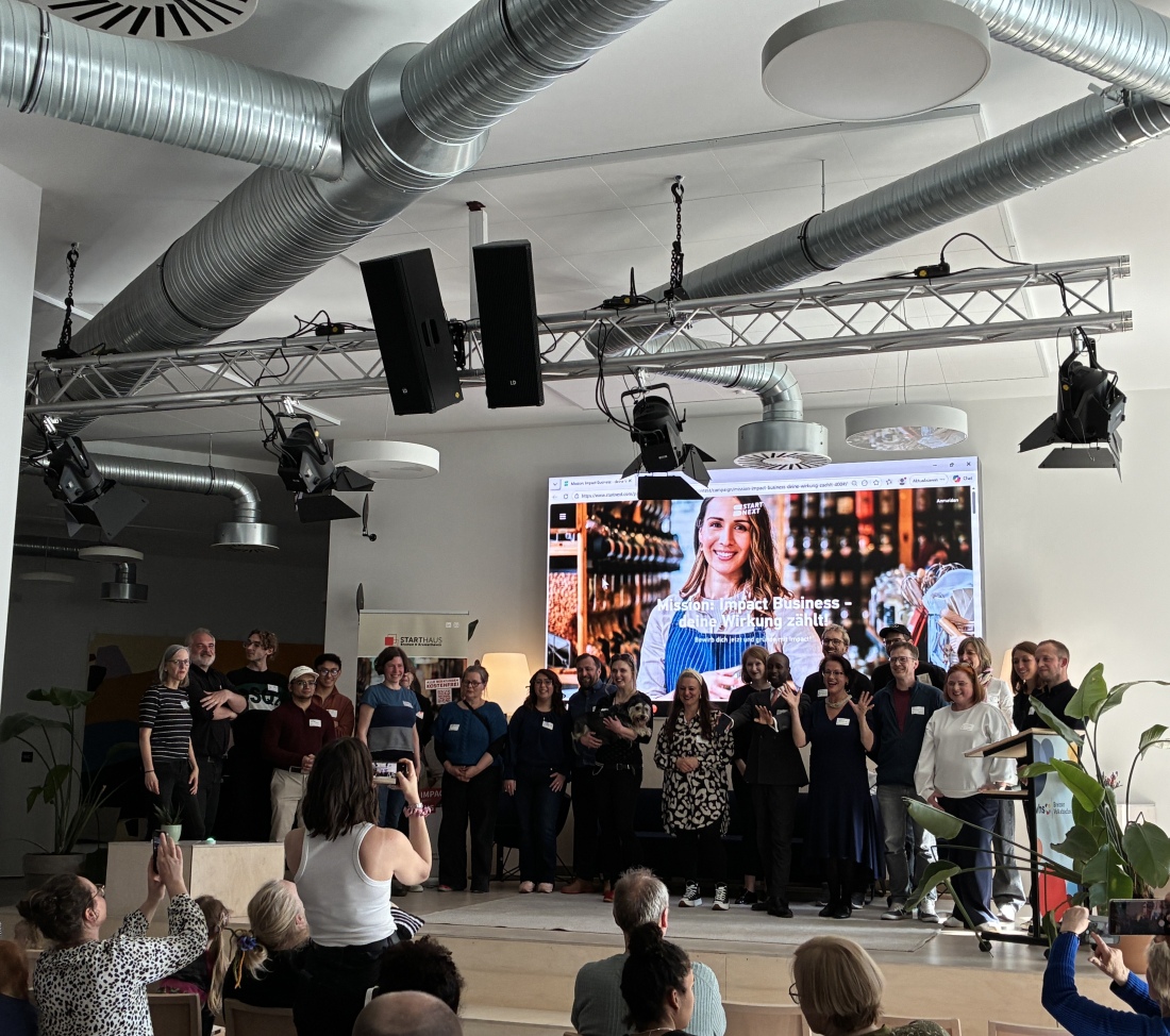 Eine Bühne, auf der alle Projekte im Rahmen des Crowdfunding-Contests "Mission: Impact Business" präsentiert werden. Auf einer Leinwand sind die Projektnamen zu sehen, während Teilnehmende aus dem Publikum klatschen.