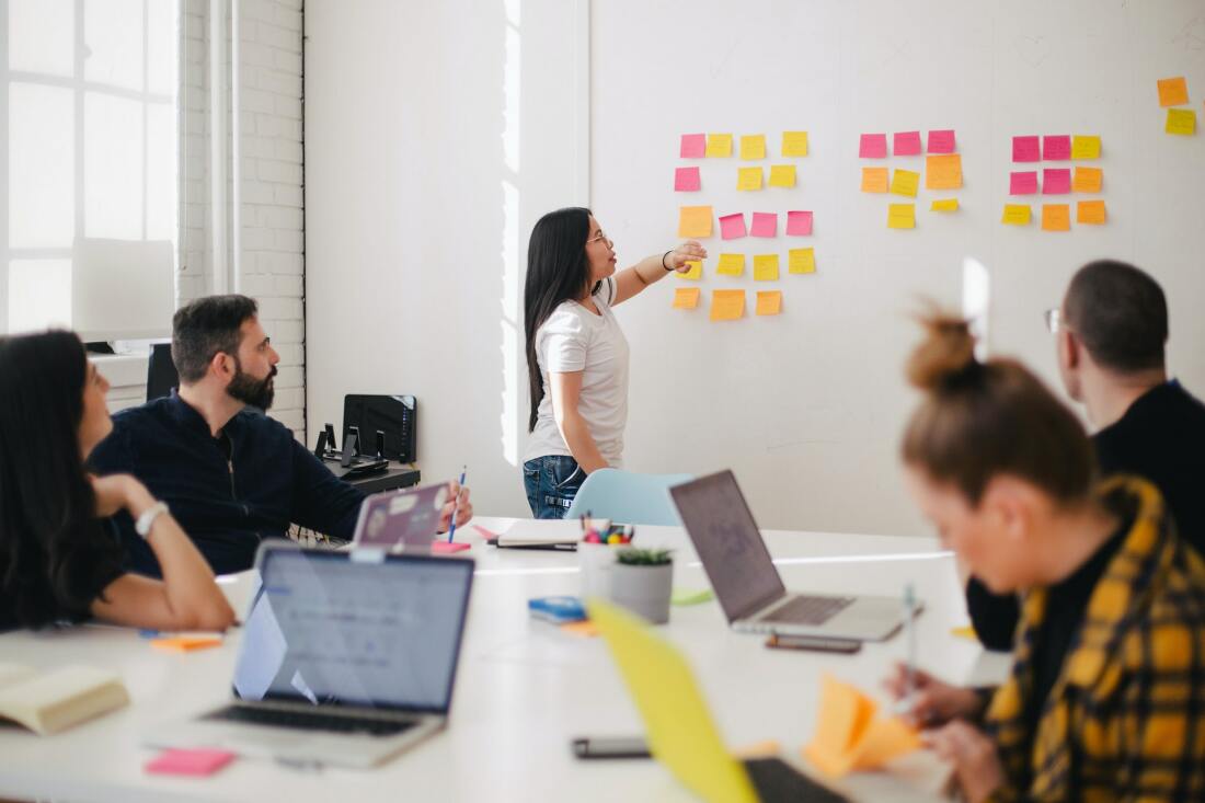 Menschen arbeiten am großen Tisch, eine Frau steht an einem White Board mit vielen bunten Post-its.